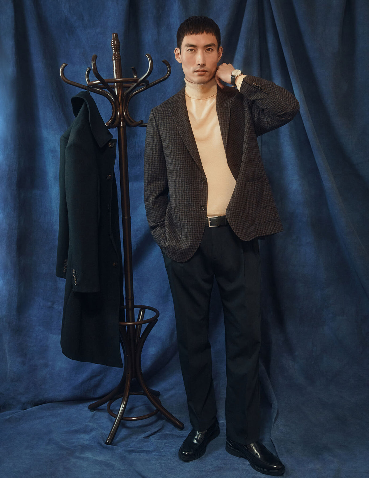A man wearing a tan turtleneck, a dark patterned blazer, and black Tech Wool Suit Pants Relaxed Fit Black stands next to a wooden coat rack holding a dark coat.