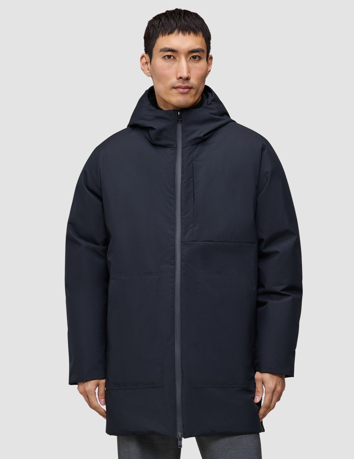 A man wears a black Utility Jacket Black with a hood that zips to the chin, featuring a vertical chest pocket and two large front pockets.