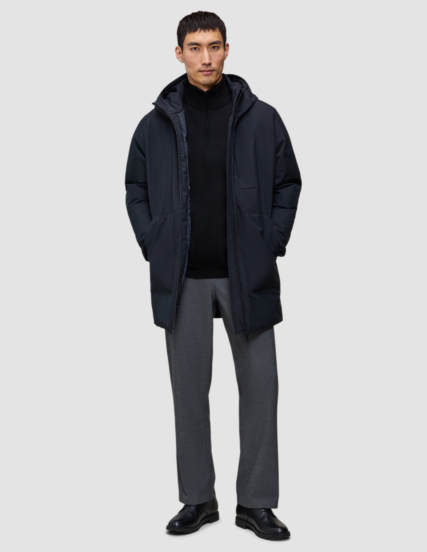 A man wearing a black Utility Jacket Black over a black zip-up sweater and grey trousers.
