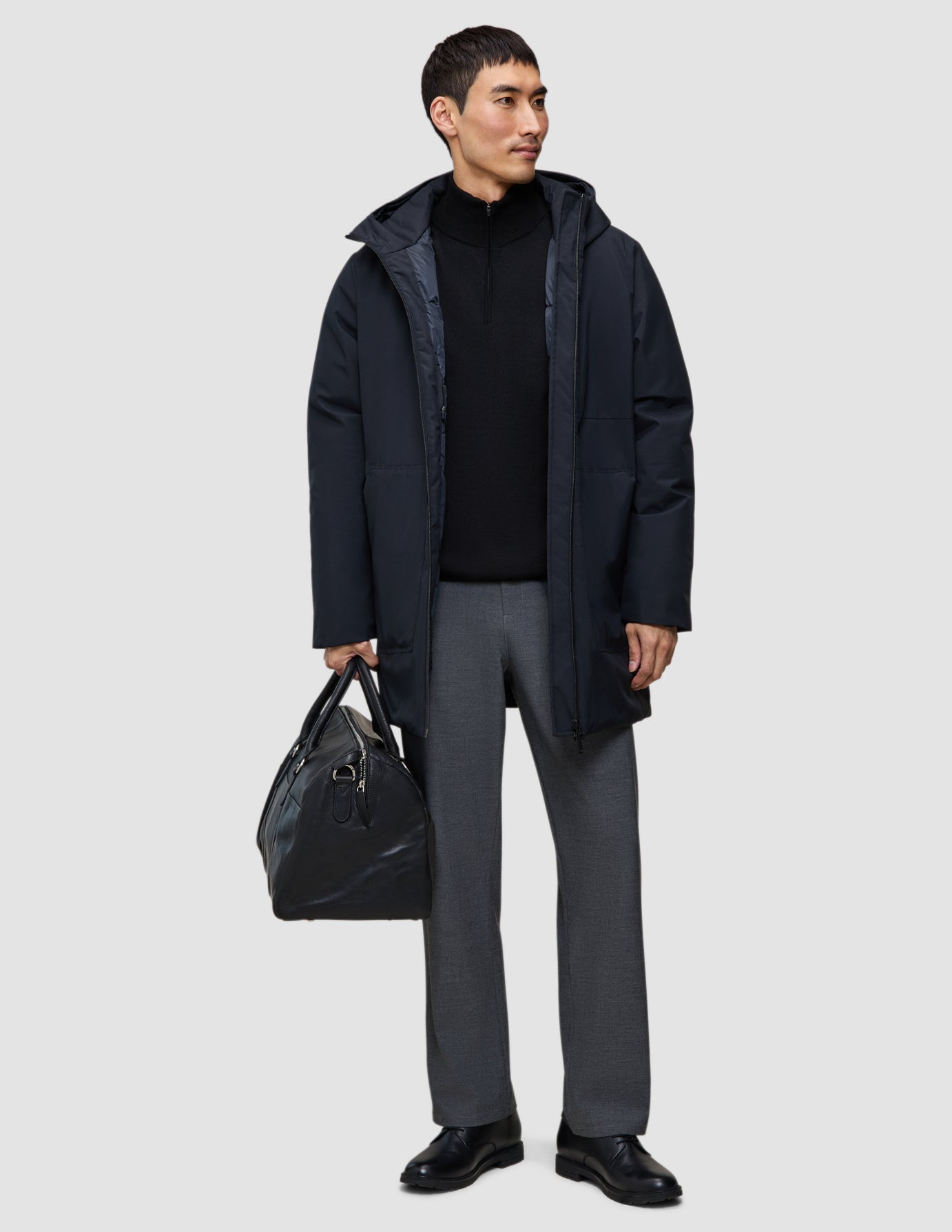 A man wears a black Utility Jacket Black hooded waterproof utility jacket, a black quarter-zip sweater, and grey trousers while holding a black leather duffel bag.