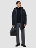 A man wears a black Utility Jacket Black hooded waterproof utility jacket, a black quarter-zip sweater, and grey trousers while holding a black leather duffel bag.