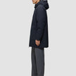 A person stands in profile wearing a Utility Jacket Black, gray trousers, and black shoes.