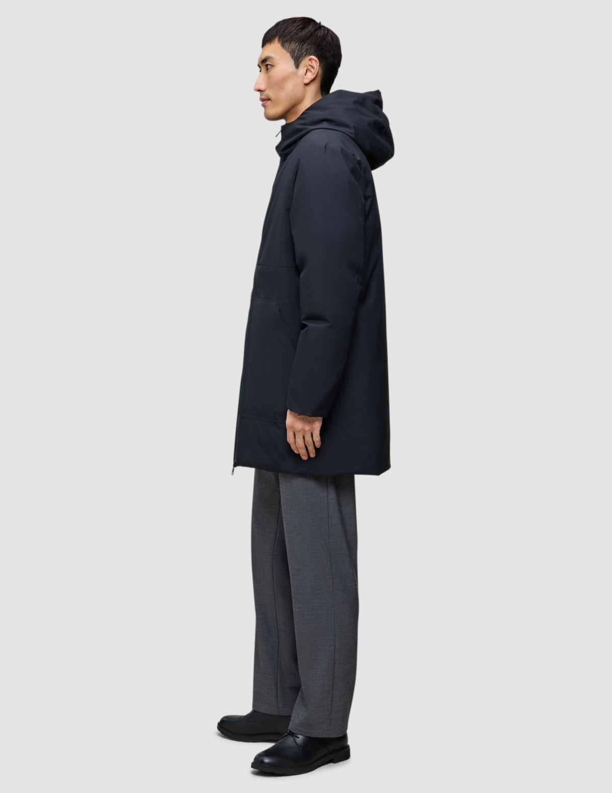 A person stands in profile wearing a Utility Jacket Black, gray trousers, and black shoes.