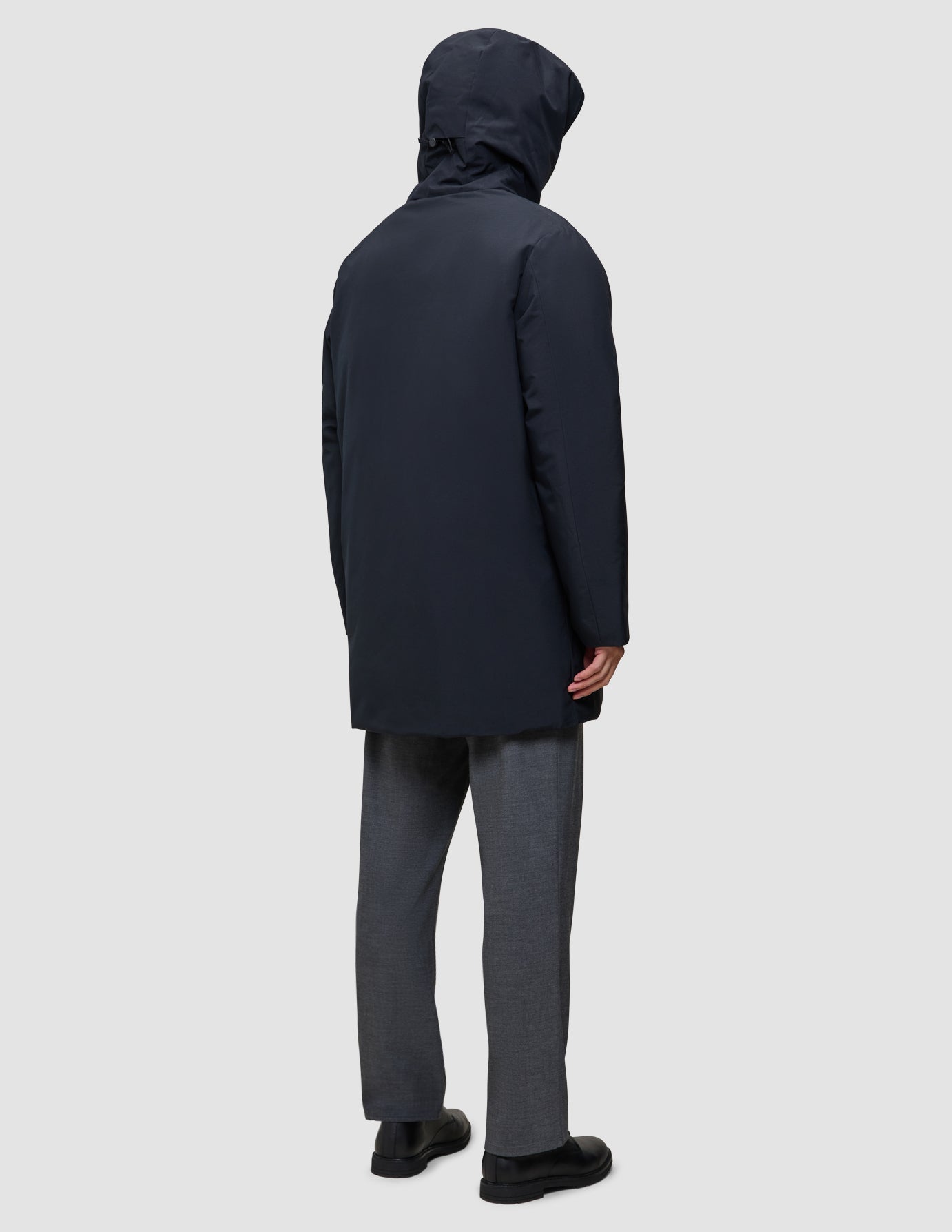 A person seen from behind wearing a black Utility Jacket Black hooded waterproof jacket, gray trousers, and black shoes.