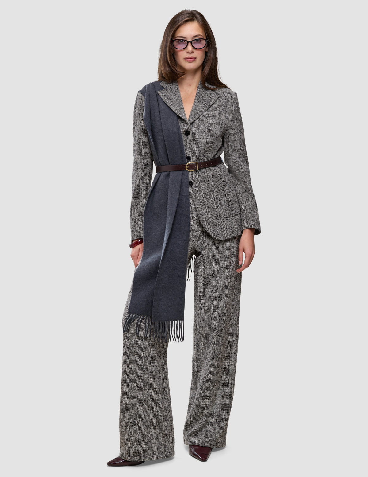 A woman wears a gray herringbone bouclé blazer, matching Women Ashryn Pants Wide Domino Herringbone, a dark blue scarf, a brown leather belt, and tinted glasses.