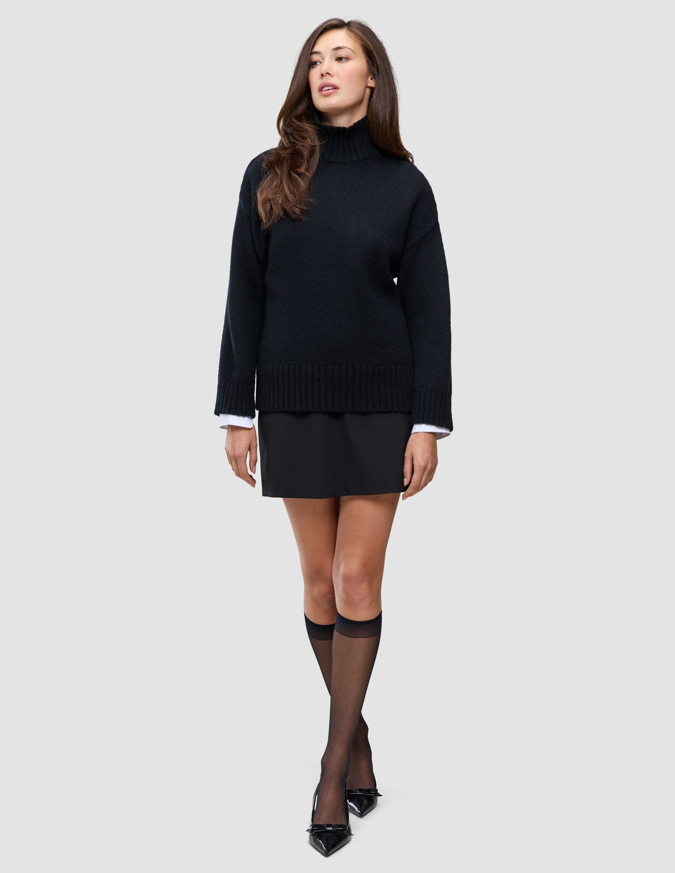 A model wearing a Women Chunky Soft Wool Knit Turtleneck Black over a white collared shirt, a black skirt, knee-high stockings, and black flat shoes.
