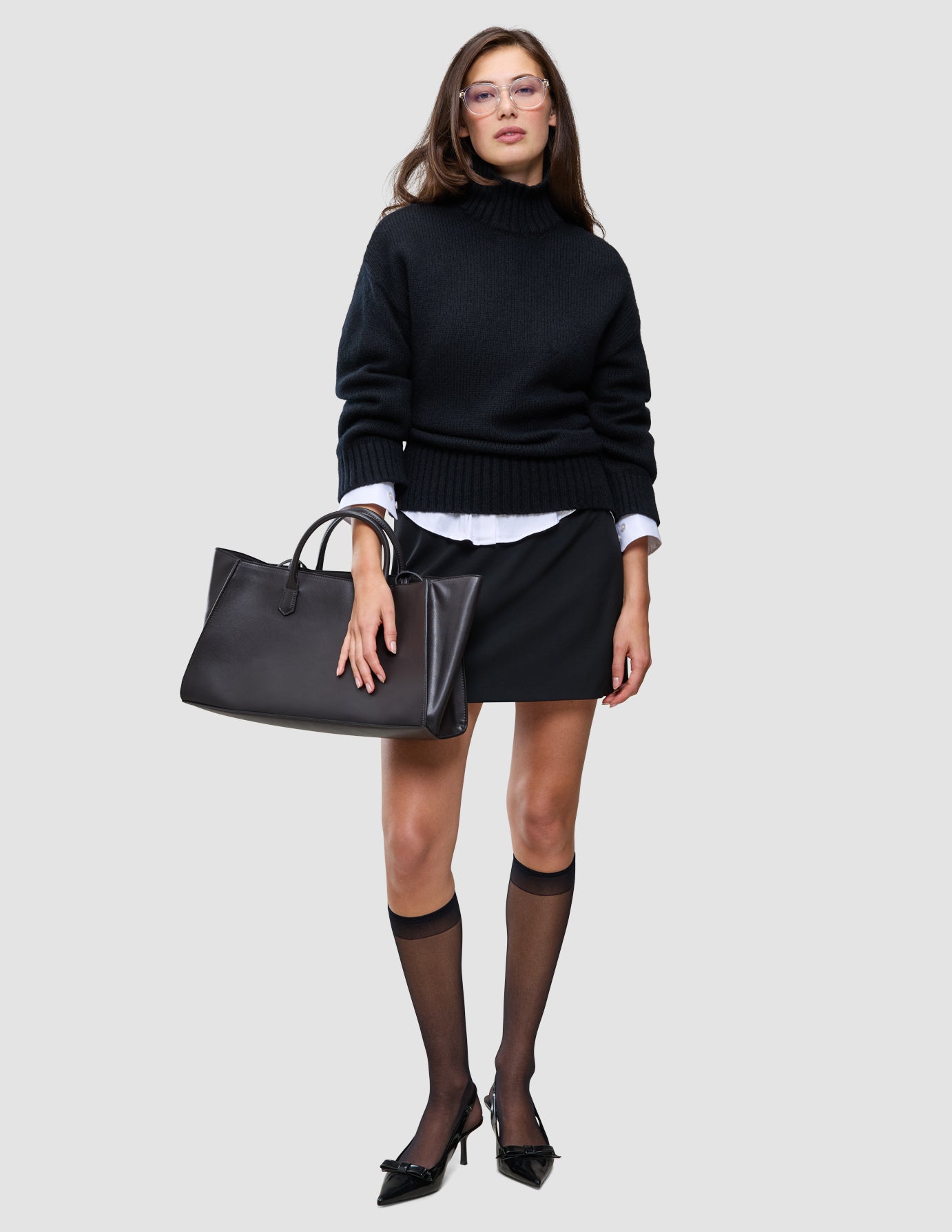 A woman wearing a Women Chunky Soft Wool Knit Turtleneck Black over a white collared shirt, a black mini skirt, sheer black stockings, and black kitten heels, holding a black tote bag.