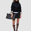 A woman wearing a Women Chunky Soft Wool Knit Turtleneck Black over a white collared shirt, a black mini skirt, sheer black stockings, and black kitten heels, holding a black tote bag.