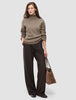 A woman wearing a Women Chunky Soft Wool Knit Turtleneck Taupe Melange sweater and dark pinstriped wide-leg trousers, holding a brown suede tote bag.