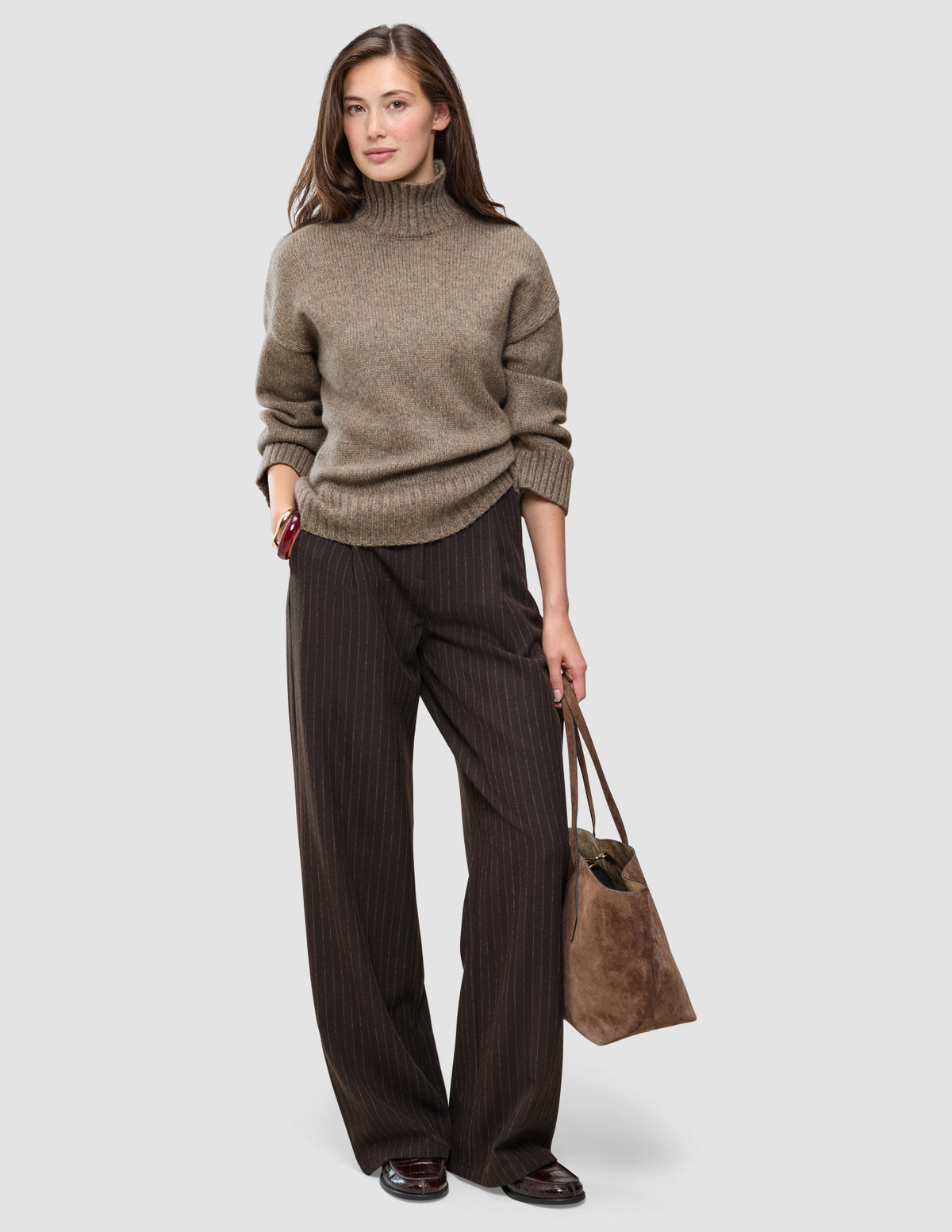 A woman wearing a Women Chunky Soft Wool Knit Turtleneck Taupe Melange sweater and dark pinstriped wide-leg trousers, holding a brown suede tote bag.