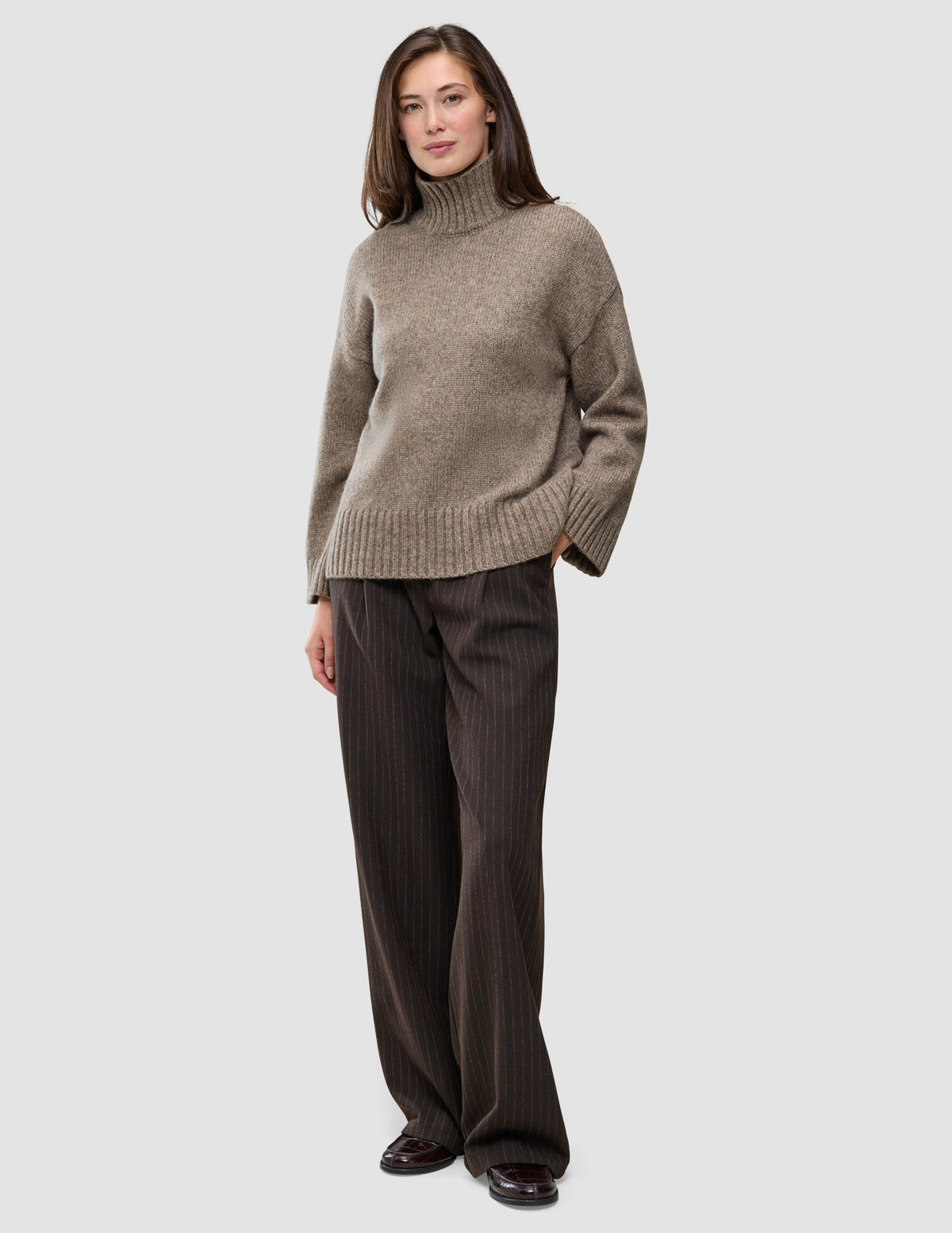 A woman wearing a Women Chunky Soft Wool Knit Turtleneck Taupe Melange in a Scandinavian design with dark brown pinstriped trousers.