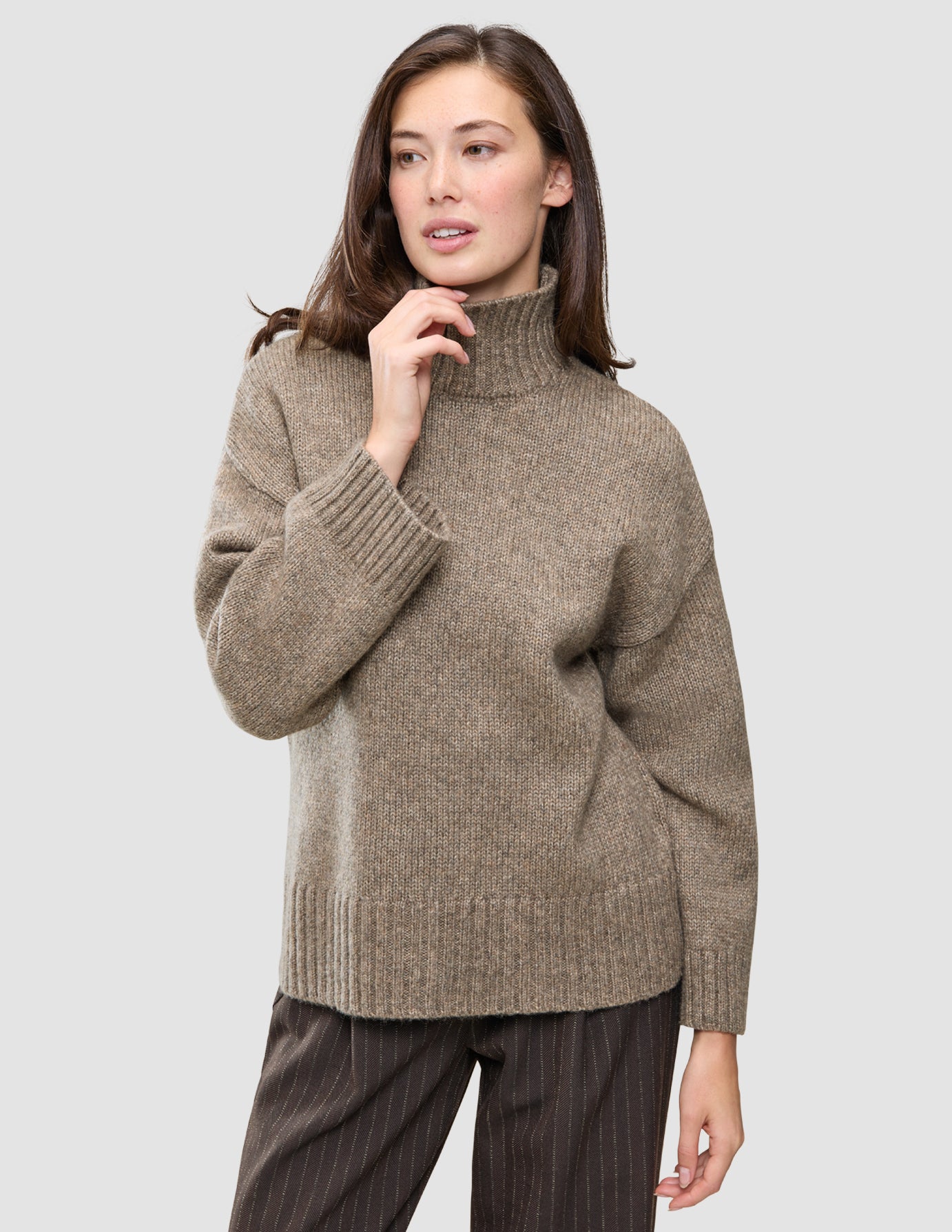 A woman wearing the Women Chunky Soft Wool Knit Turtleneck Taupe Melange, featuring a taupe, chunky knit merino wool turtleneck with ribbed cuffs and hem.