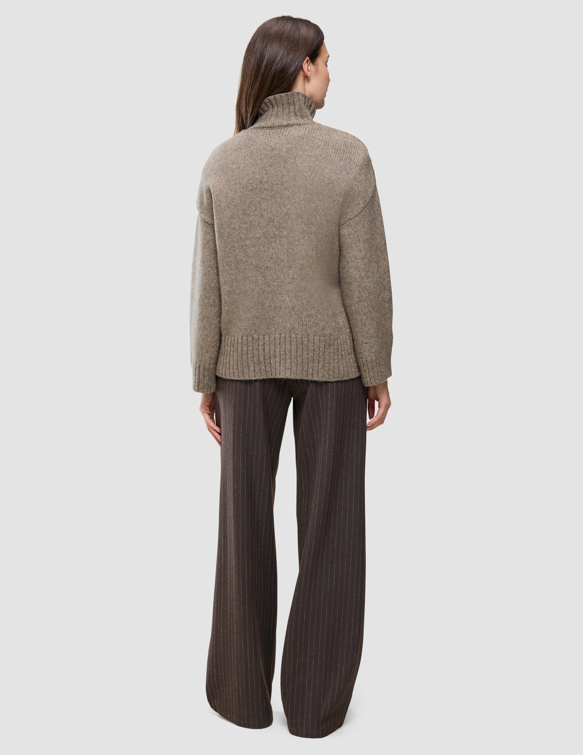 Seen from behind, a person wears the Women Chunky Soft Wool Knit Turtleneck Taupe Melange paired with dark brown pinstriped trousers, reflecting a minimalist Scandinavian design.