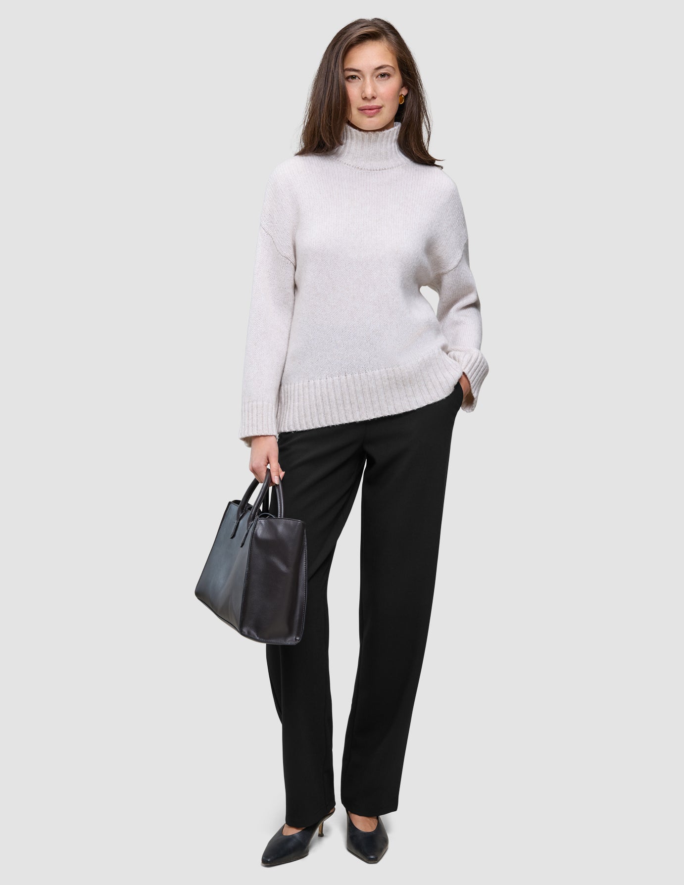 A woman wearing a beige turtleneck sweater and Women GO2 Pants Straight Black, holding a black handbag.