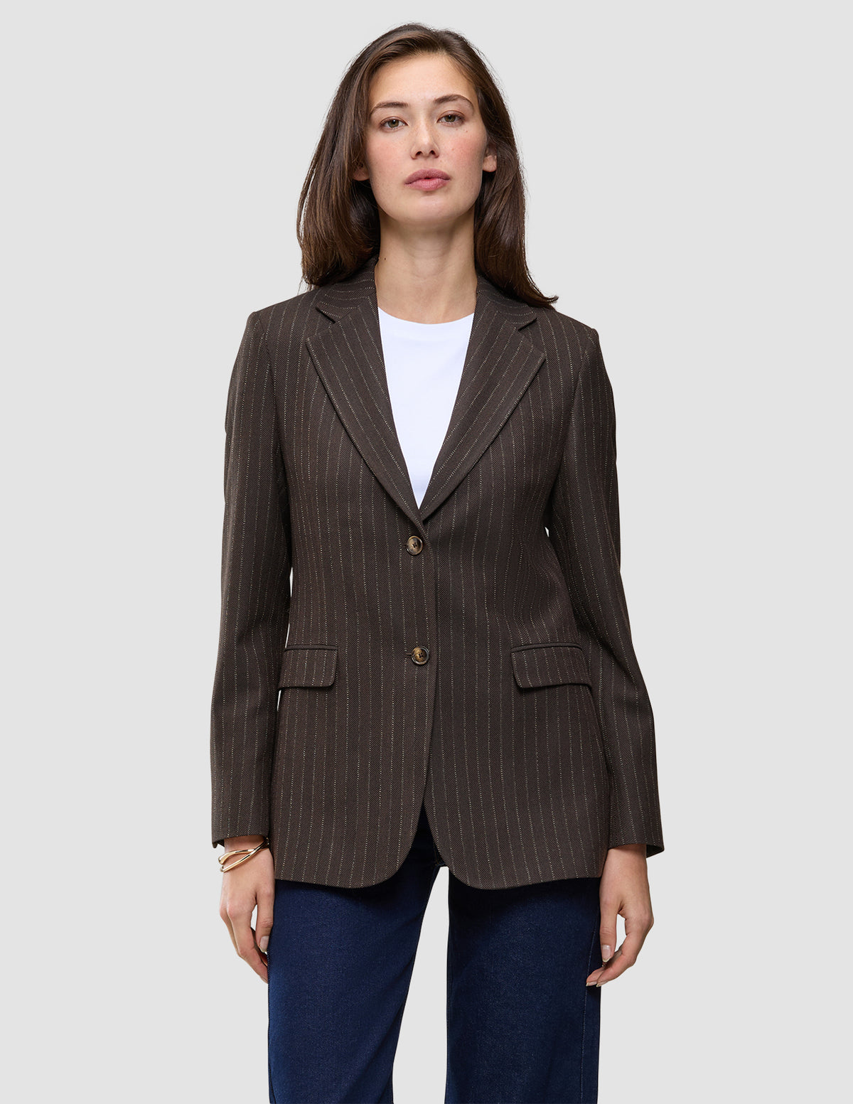 A woman wears a brown Women Serene Blazer Regular Walnut Herringbone with a faint pinstripe pattern, a white t-shirt, and blue jeans, reflecting a clean Scandinavian design.
