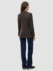 Back view of a woman wearing a Women Serene Blazer Regular Walnut Herringbone, dark blue jeans, and brown shoes.