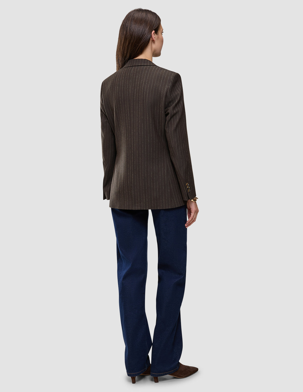 Back view of a woman wearing a Women Serene Blazer Regular Walnut Herringbone, dark blue jeans, and brown shoes.