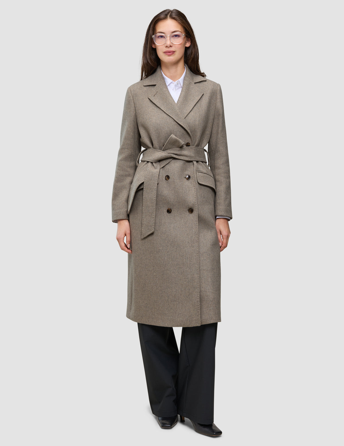 A woman wears a Women Wool Coat Greige, a double-breasted, knee-length grey wool coat with a belted waist over a white shirt and black trousers.