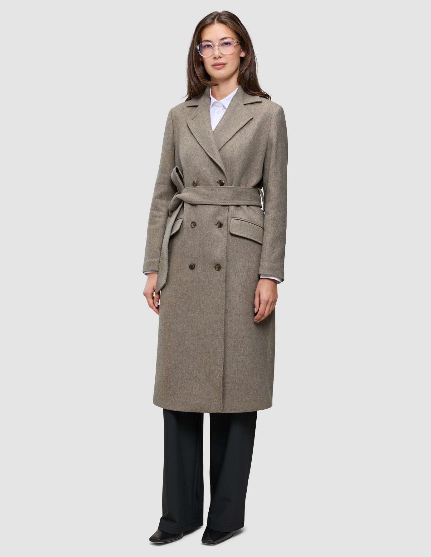 A woman wears a Women Wool Coat Greige, a long, double-breasted grey-brown wool coat with a matching tie belt over a white shirt and black trousers, showcasing minimalist Scandinavian design.