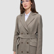 A person wears a Women Wool Coat Greige, featuring a light brown-gray, double-breasted wool design with a belt over a white collared shirt.