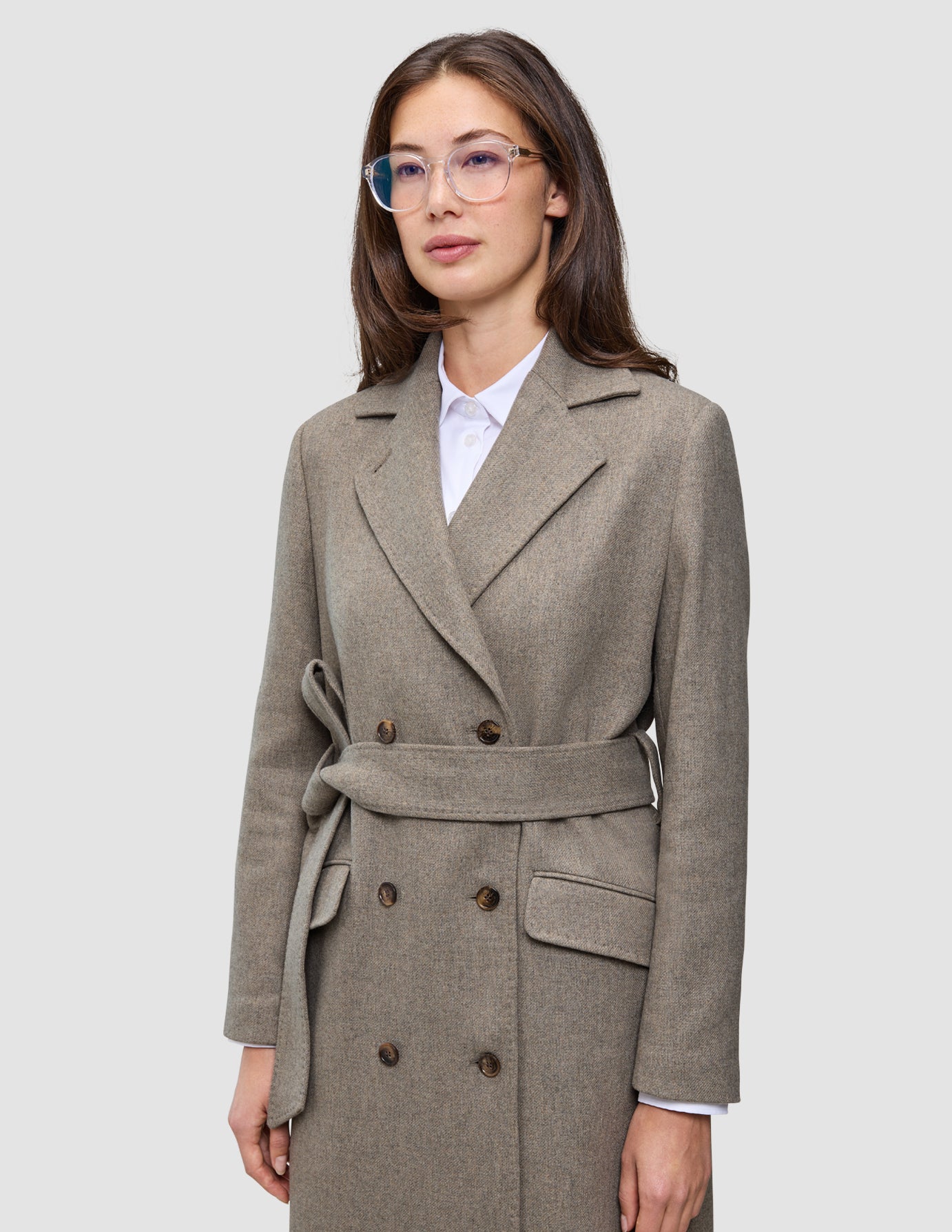 A person wears a Women Wool Coat Greige, featuring a light brown-gray, double-breasted wool design with a belt over a white collared shirt.