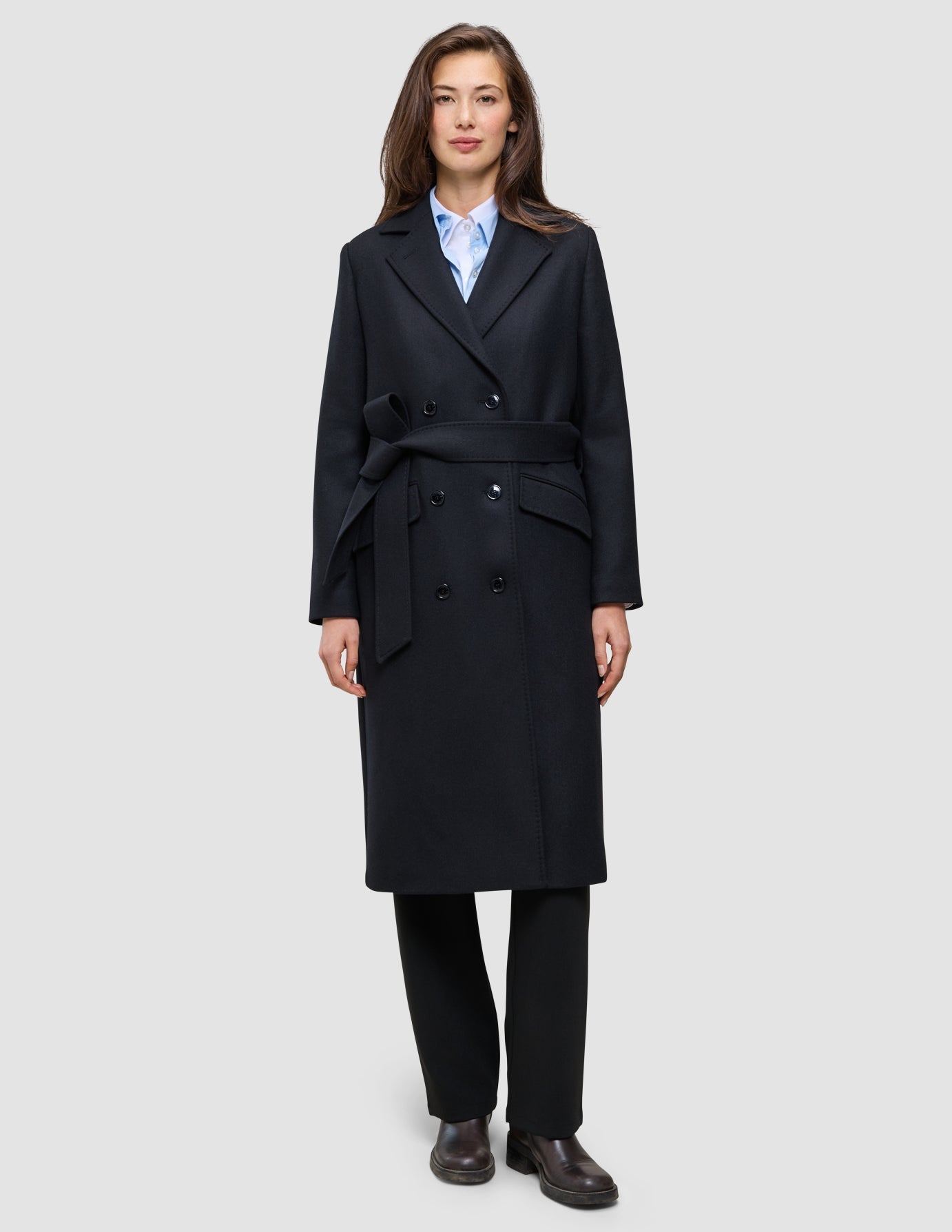 A woman wears a handmade-in-Turkey Women’s Wool Coat Black in a double-breasted style with a belted waist over a light blue collared shirt and black trousers.
