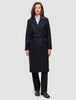 A woman wears a handmade-in-Turkey Women’s Wool Coat Black in a double-breasted style with a belted waist over a light blue collared shirt and black trousers.
