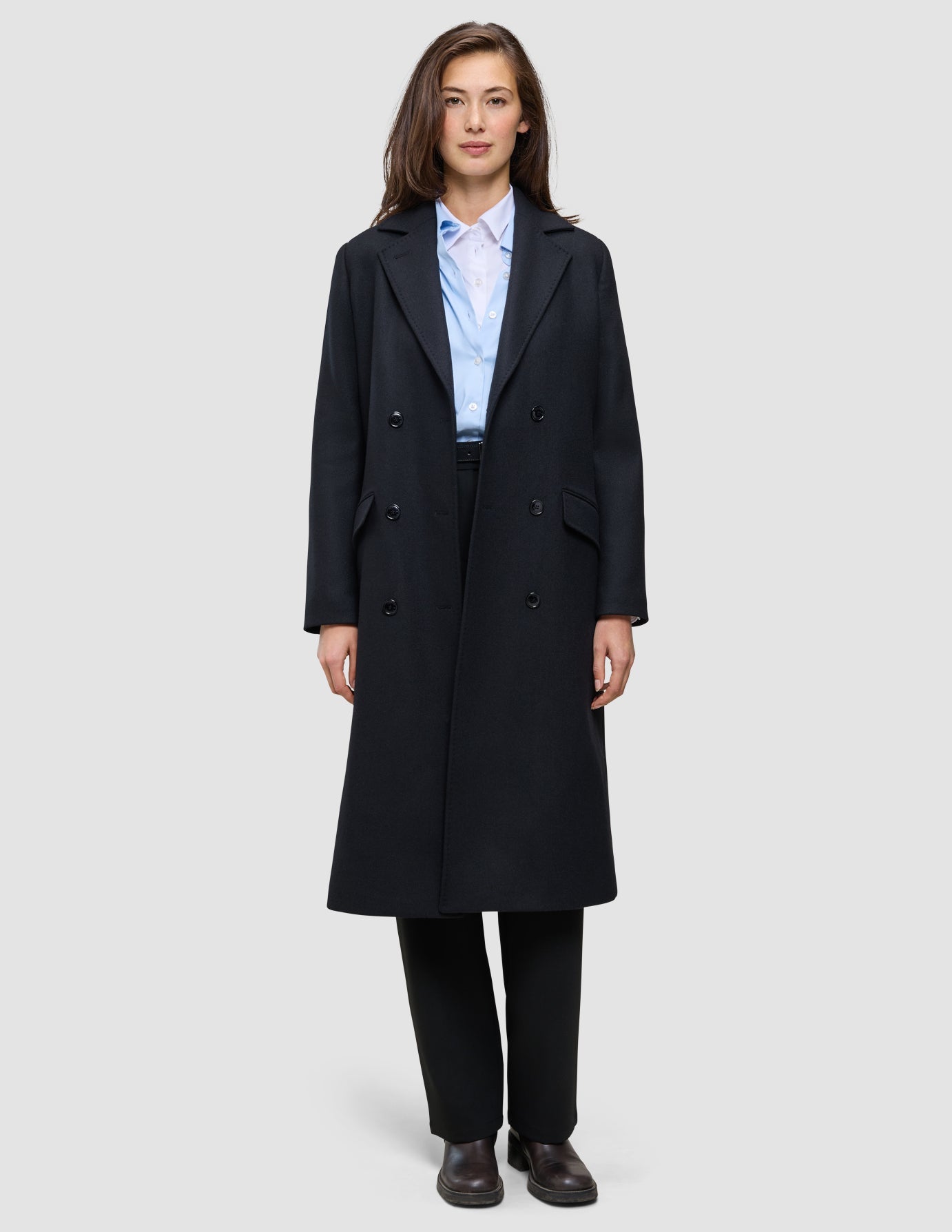 A person wearing a Women’s Wool Coat Black in a double-breasted Scandinavian design over a light blue collared shirt and black trousers.