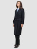 A woman wearing a Women’s Wool Coat Black in a Scandinavian-design double-breasted style with a tie waist over a light blue shirt and black trousers.