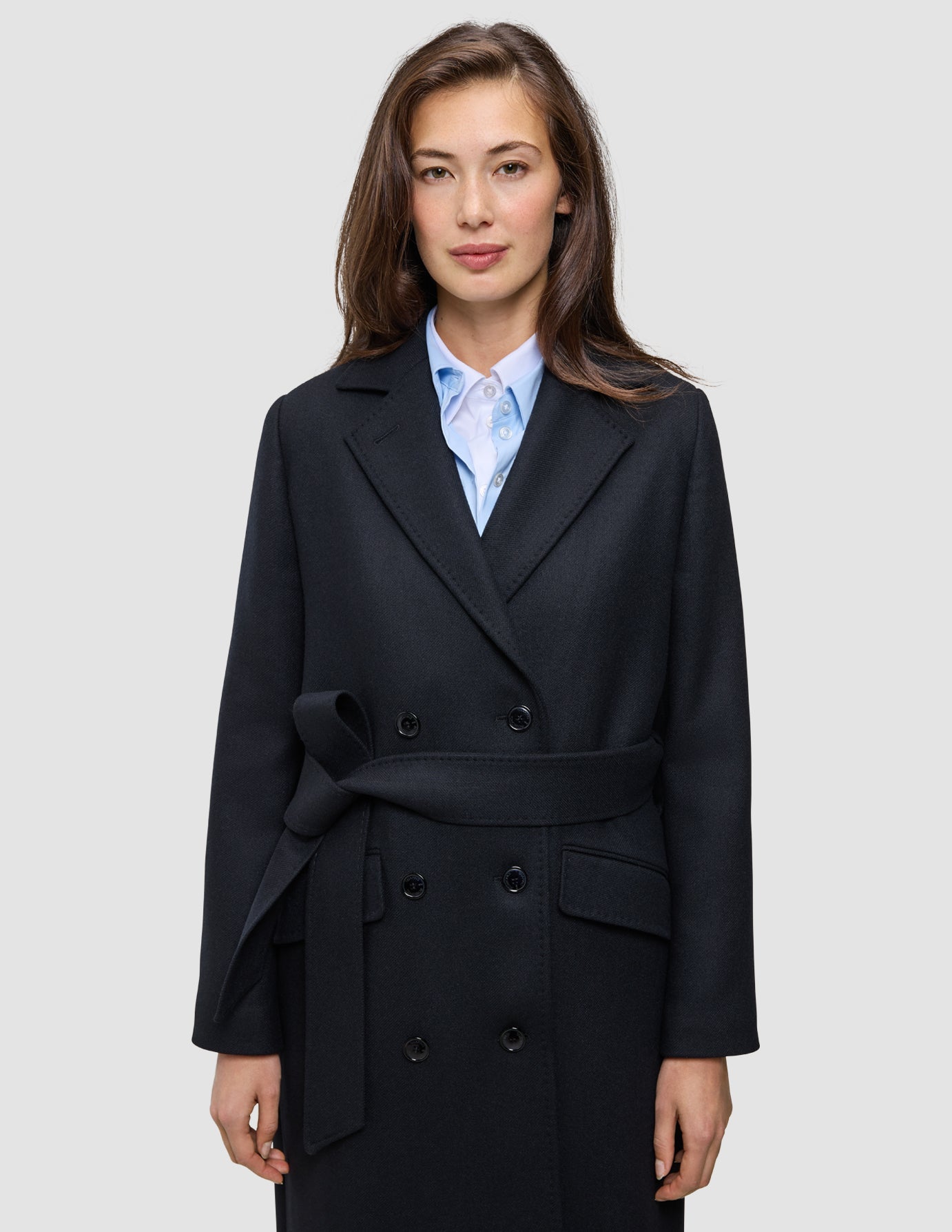 A woman wearing a Women’s Wool Coat Black in a Scandinavian-design double-breasted style with a tie waist over a light blue collared shirt.